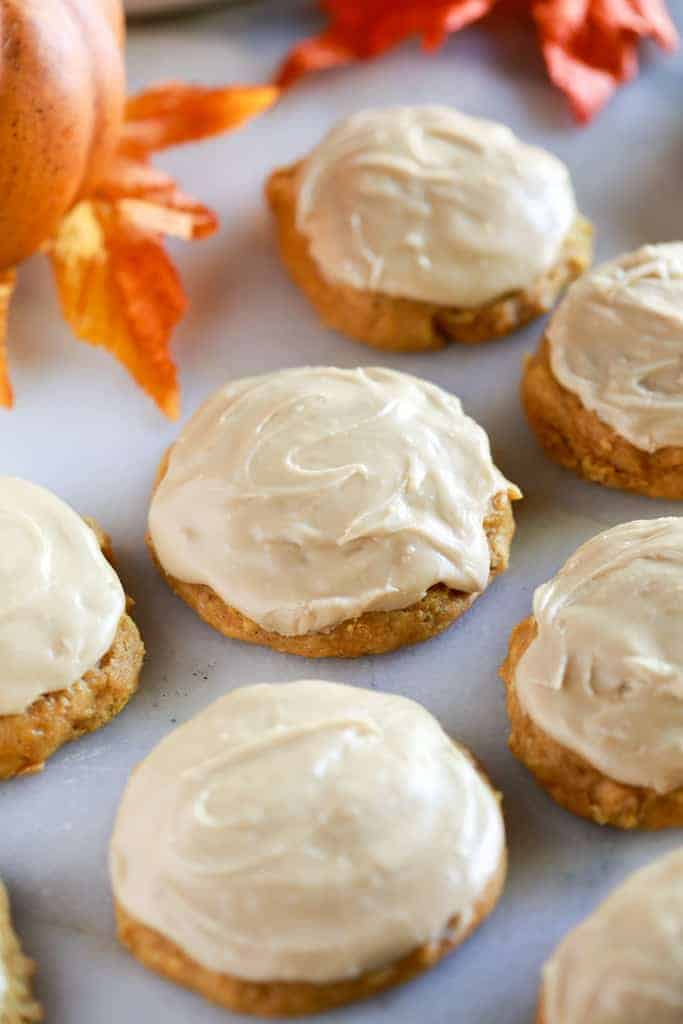 Pumpkin cookies with caramel frosting on a white marble board with fall leaves in the background.