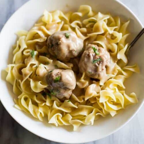Three Swedish Meatballs and gravy served over egg noodles in a white bowl with a fork.