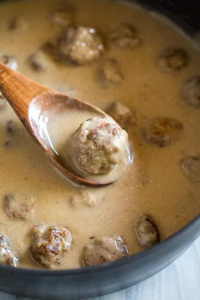 Swedish meatballs in a large skillet with gravy and a wooden spoon lifting a meatball from the pan.