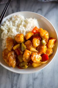 A white plate with homemade sweet and sour chicken and white rice, and chopsticks resting on the plate.