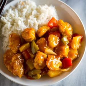 A white plate with homemade sweet and sour chicken and white rice, and chopsticks resting on the plate.