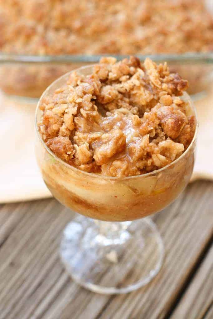 A glass goblet glass filled with apple crisp with a crumble oat topping and a pan of apple crisp in the background.