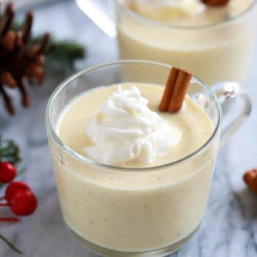 A cup of homemade eggnog served with whipped cream on top and a cinnamon stick, with holly berries, pinecones and another glass of eggnog in the background.