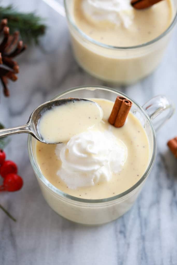 Overhead photo of two cups filled with eggnog with whipped cream and a cinnamon stick and a spoon lifting a spoonful of eggnog from the cup.