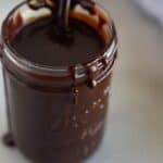 A glass mason jar filled with hot fudge sauce and a spoon in it.