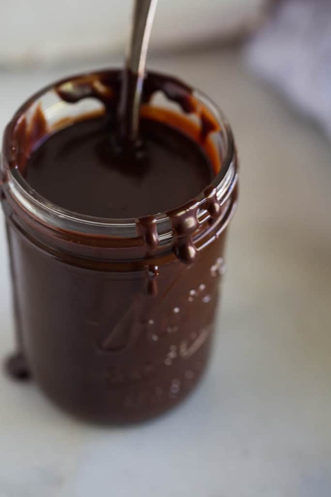 A glass mason jar filled with hot fudge sauce and a spoon in it.