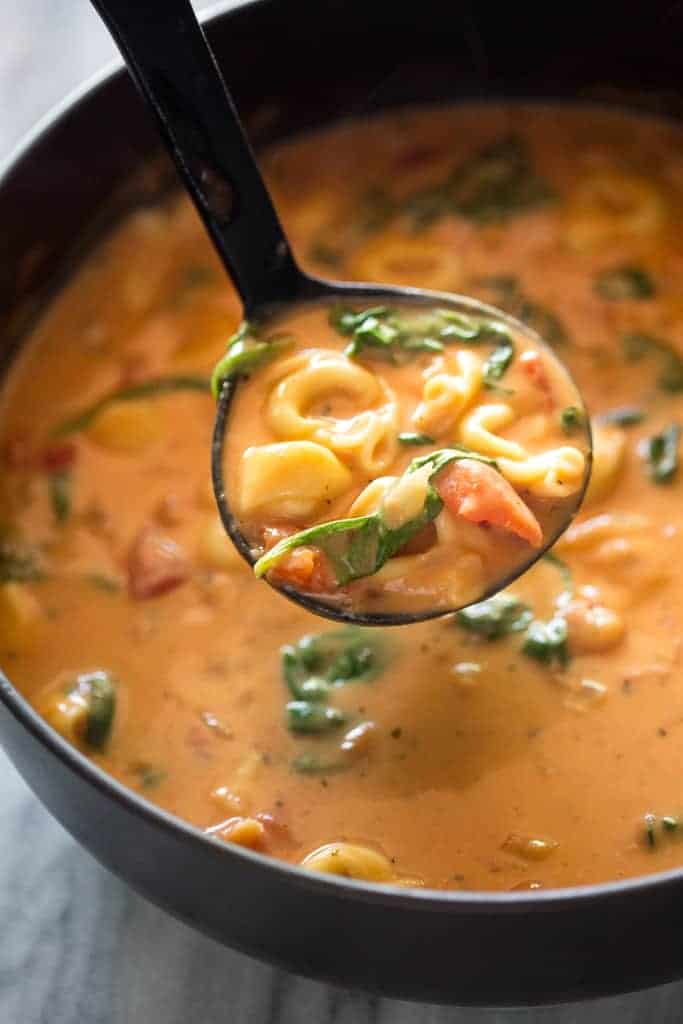 A saucepan with creamy tomato and spinach tortellini soup in it and a laddle spooning some out.