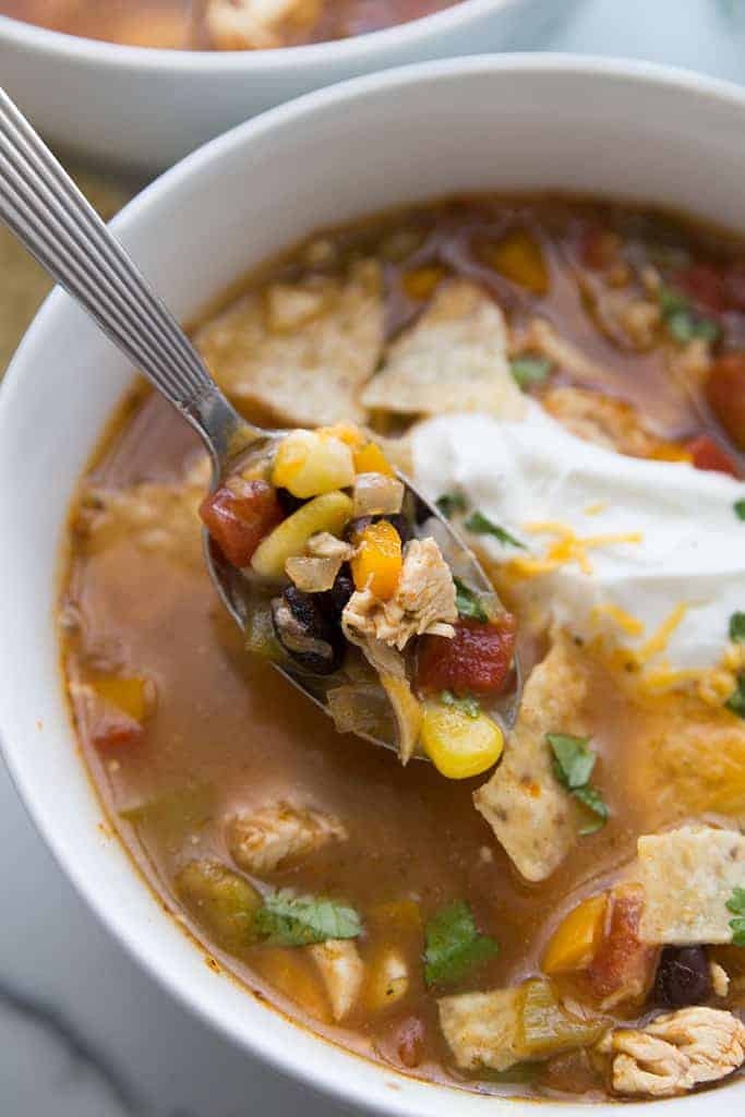 A spoon lifting chicken tortilla soup with pieces of chicken, corn, carrot, and diced tomatoes out of a white bowl