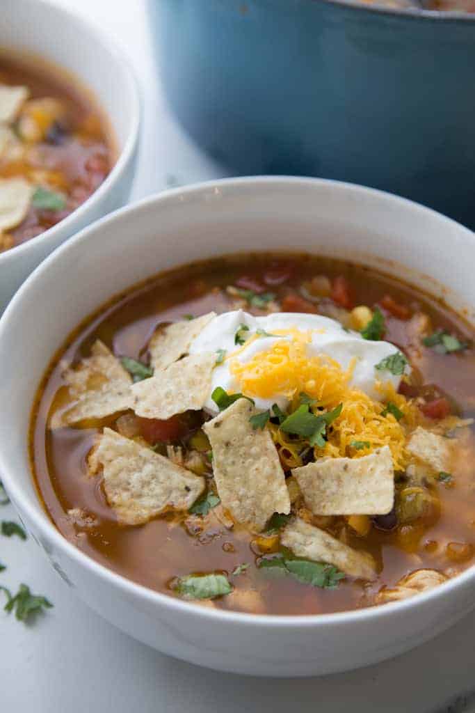 A white bowl with chicken tortilla soup in it topped with tortilla chips, sour cream, shredded cheese and cilantro.