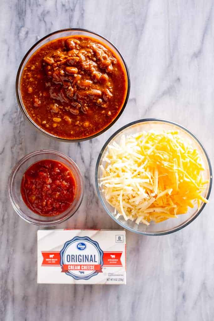 Ingredients for chili cheese dip including chili, shredded cheese, salsa and cream cheese. 