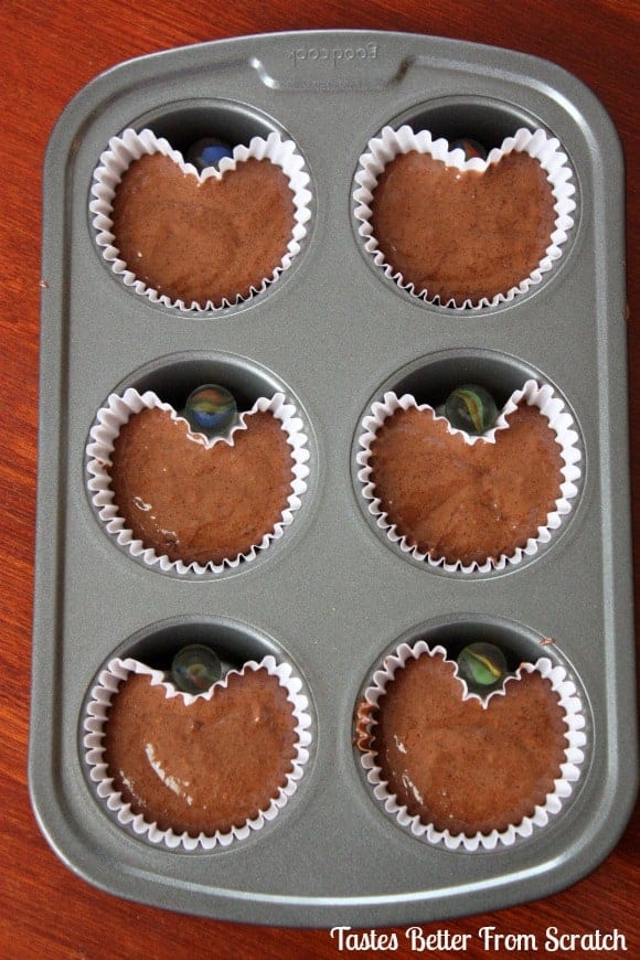 Muffin tin lined with chocolate cupcake batter in paper liners with a marble in each tin to form a heart shaped cupcake.
