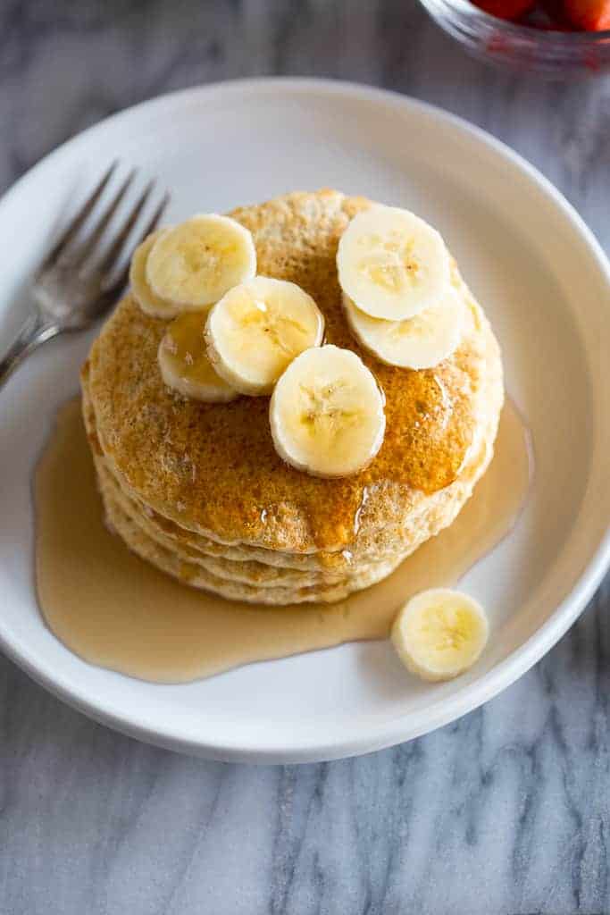 A stack of Protein Pancakes served with sliced bananas and syrup on a white plate with a fork.