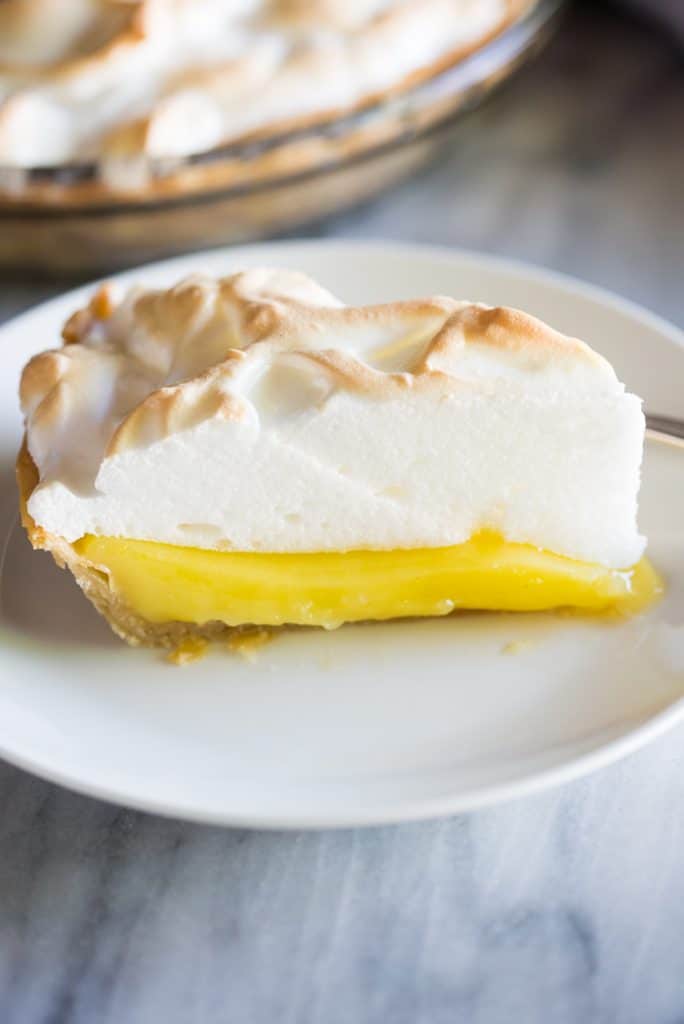 A slice of Lemon Meringue Pie served on a white plate with the remaining pie in the background.