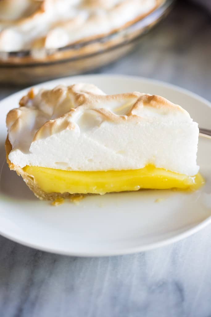 A slice of Lemon Meringue Pie served on a white plate with the remaining pie in the background.