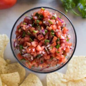 Overhead photo of pico de gallo served in a clear bowl, with tortilla chips, cilantro, lime and tomatoes in the background.