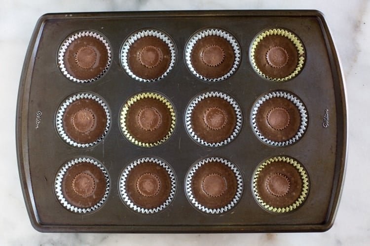 A cupcake pan with liners filled with chocolate cupcake batter and a mini Reese's chocolate in the center of each cupcake, ready to bake.