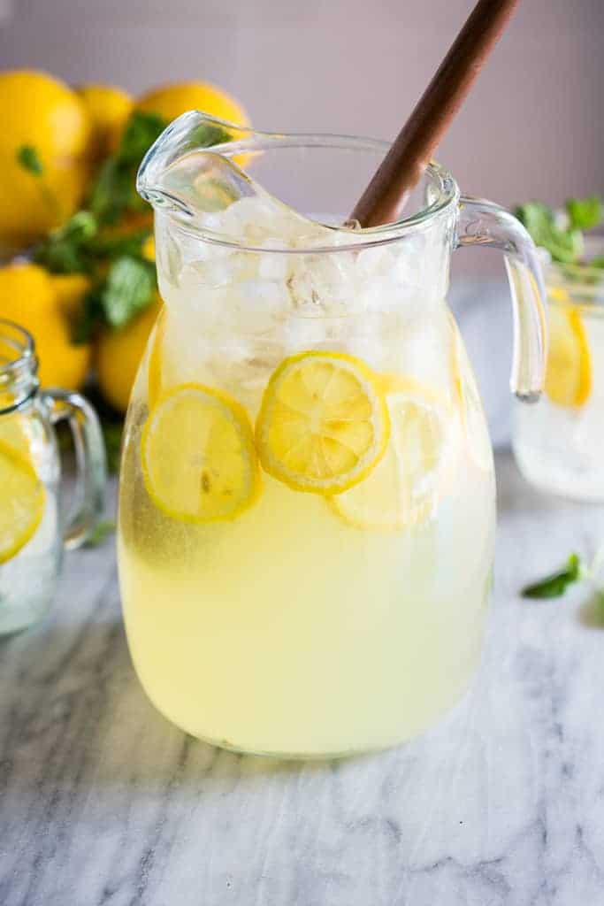 A pitcher of fresh squeezed lemonade with lemon slices and ice and lemons in the background.