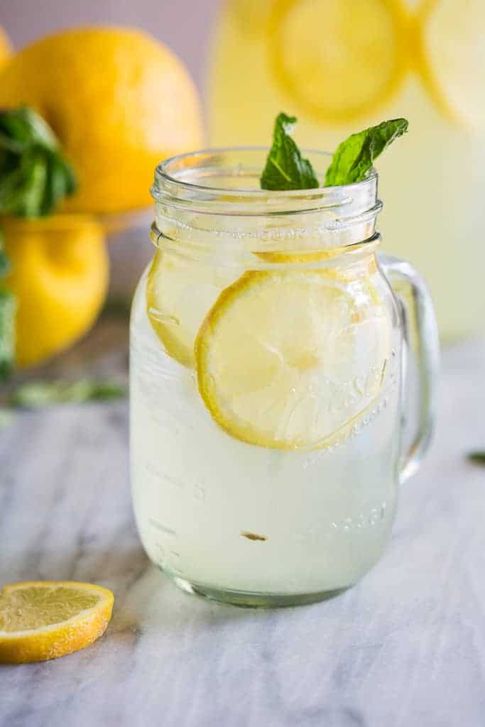 A mason jar with homemade lemonade, ice, lemon slices and mint and more whole lemons and mint in the background.