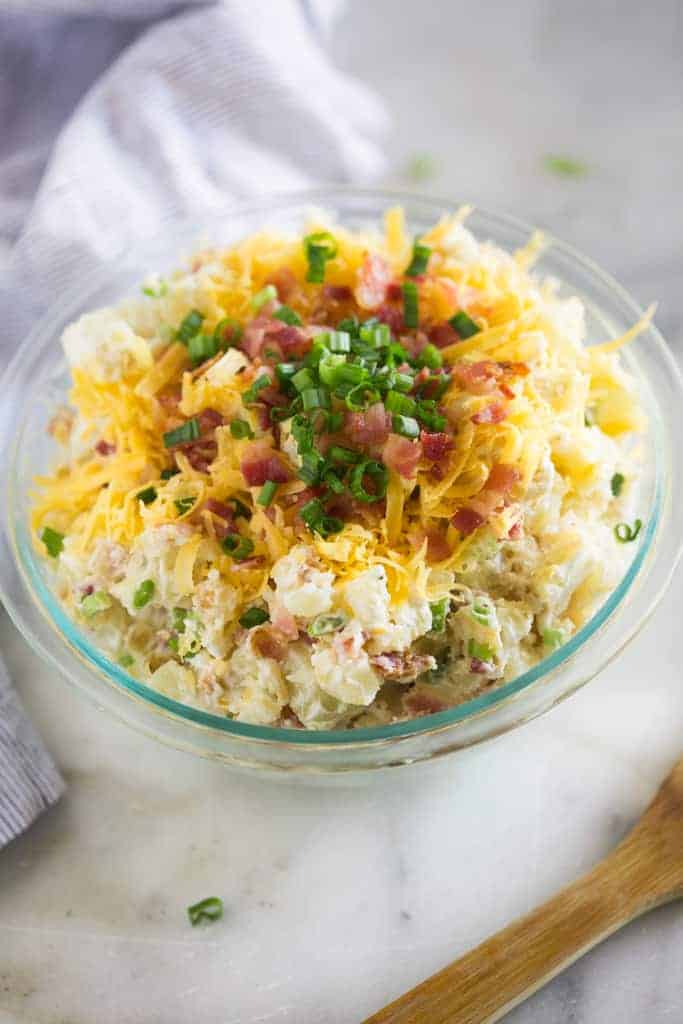 Baked potato salad served in a glass bowl topped with a pile of shredded cheddar cheese, chopped bacon and green onions.