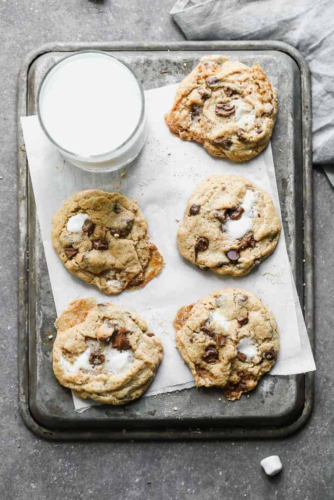 Five cookies on parchment paper on a baking sheet, next to a glass of milk.