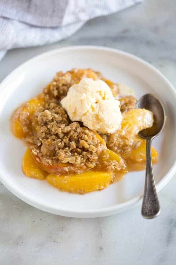 Peach crisp served with a scoop of vanilla ice cream on top, on a white plate with a spoon.