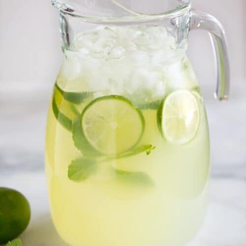 A clear glass pitcher filled with mint limeade, ice, slices of fresh lime and springs of fresh mint, ready to serve.