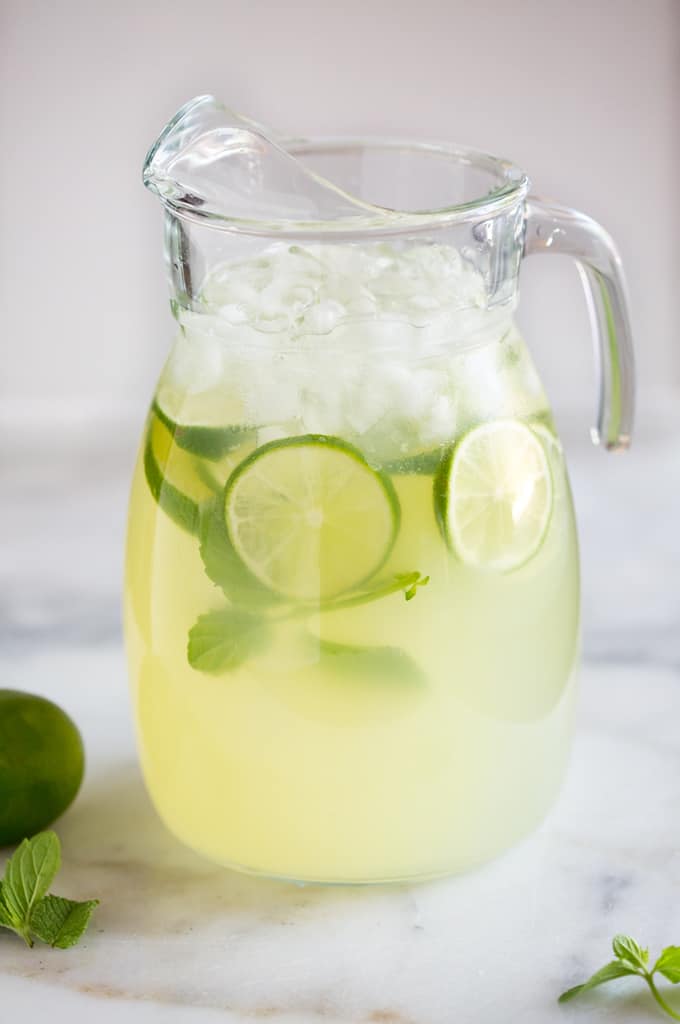 A clear glass pitcher filled with mint limeade, ice, slices of fresh lime and springs of fresh mint, ready to serve.