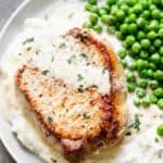 A white plate with mashed potatoes, a pork chop and creamy mustard sauce on top and peas on the side.