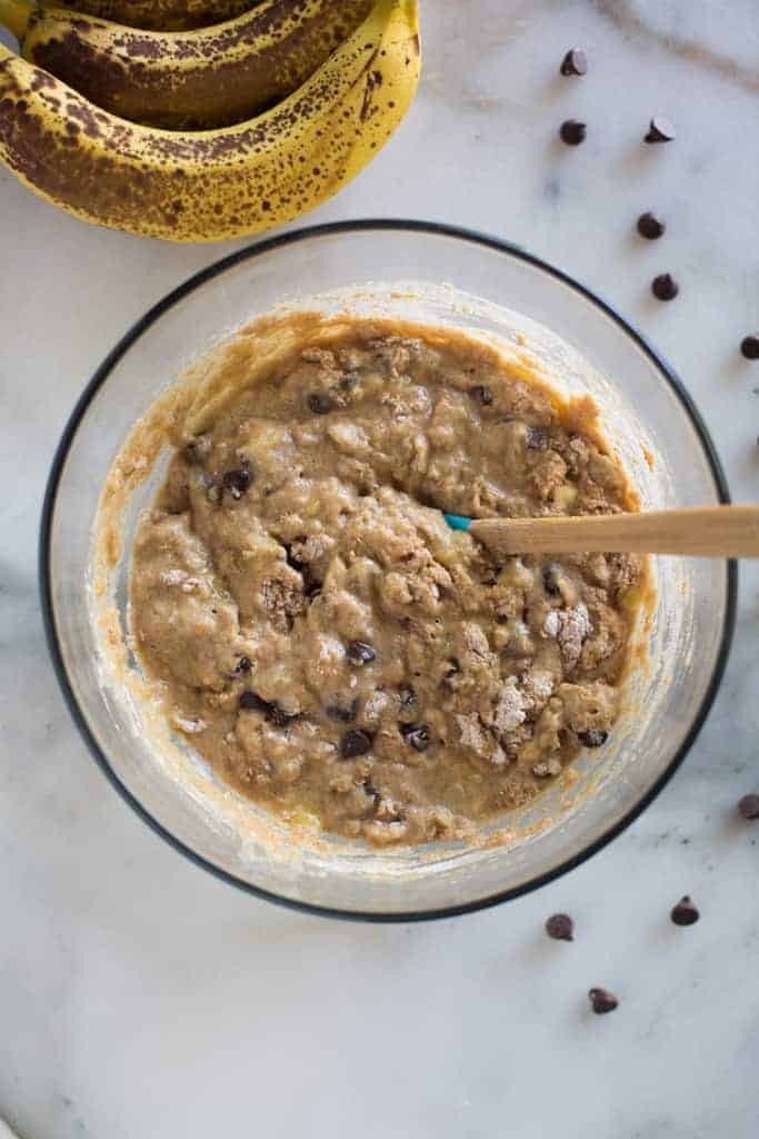 Whole wheat pancake batter with mashed bananas and chocolate chips in it, in a glass bowl with a spatula to stir it and overripe bananas and chocolate chips in the background.