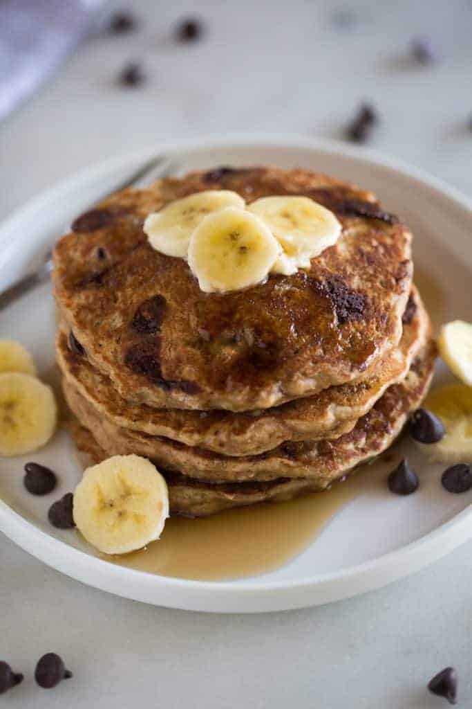 A stack of whole wheat banana bread pancakes on a white plate topped with sliced bananas with chocolate chips on the plate and in the background.