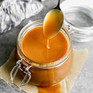 A spoon dripping caramel sauce into a glass jar, with some of the sauce pouring over the side of the jar.