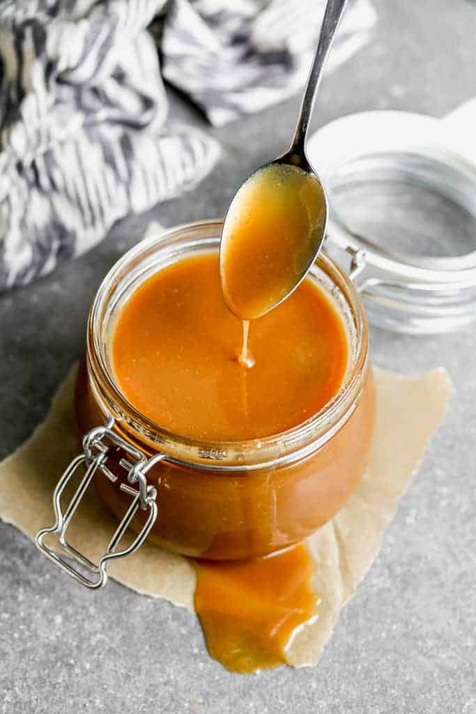 A spoon dripping caramel sauce into a glass jar full and some caramel spilling over the side of the jar.