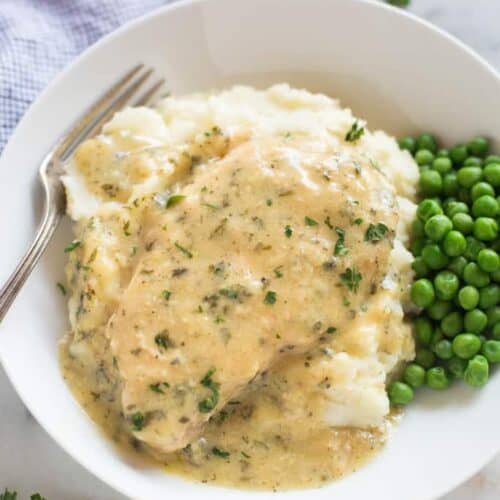 Slow cooker cream ranch pork chops served in a white plate over a bed of mashed potatoes, with extra sauce and a side of green peas.