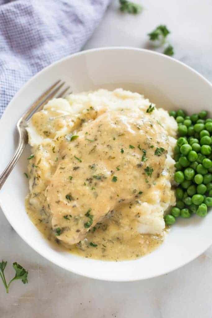 Slow cooker cream ranch pork chops served in a white plate over a bed of mashed potatoes, with extra sauce and a side of green peas.
