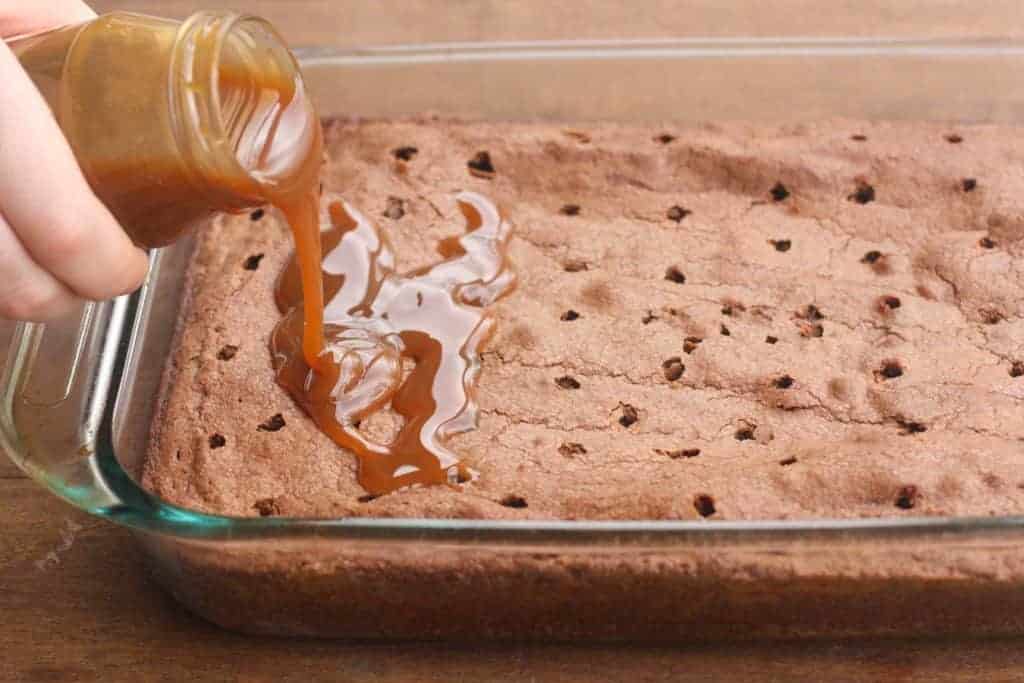 A 9x13 pan of brownies with wholes poked in them and caramel sauce being poured on top.