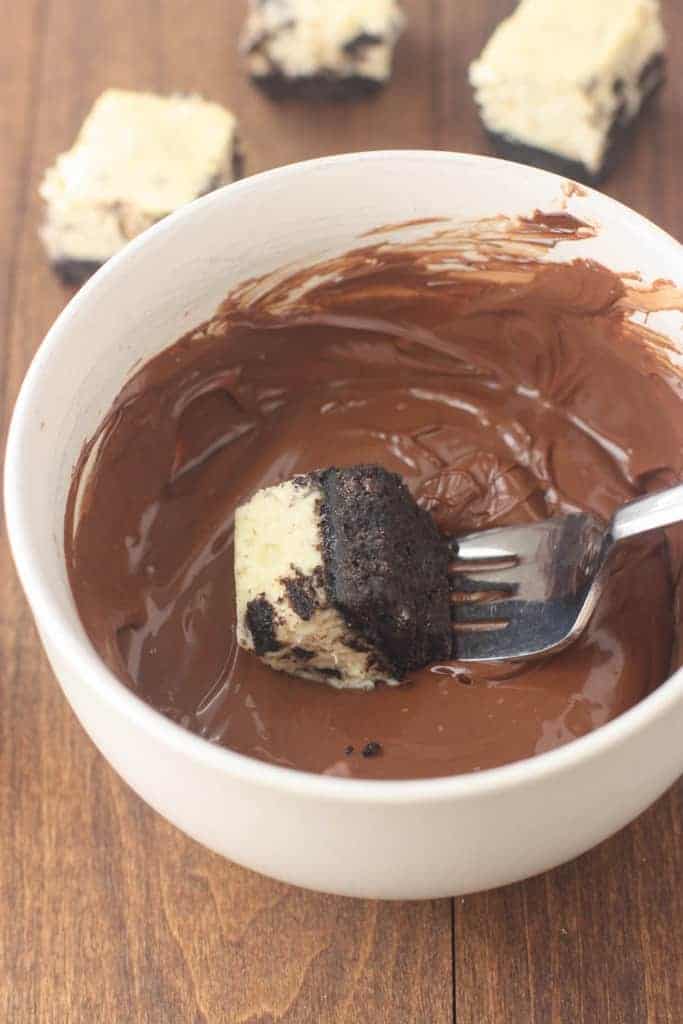 A small cheesecake bite with an Oreo crust that is being dipped in a white bowl of melted chocolate.
