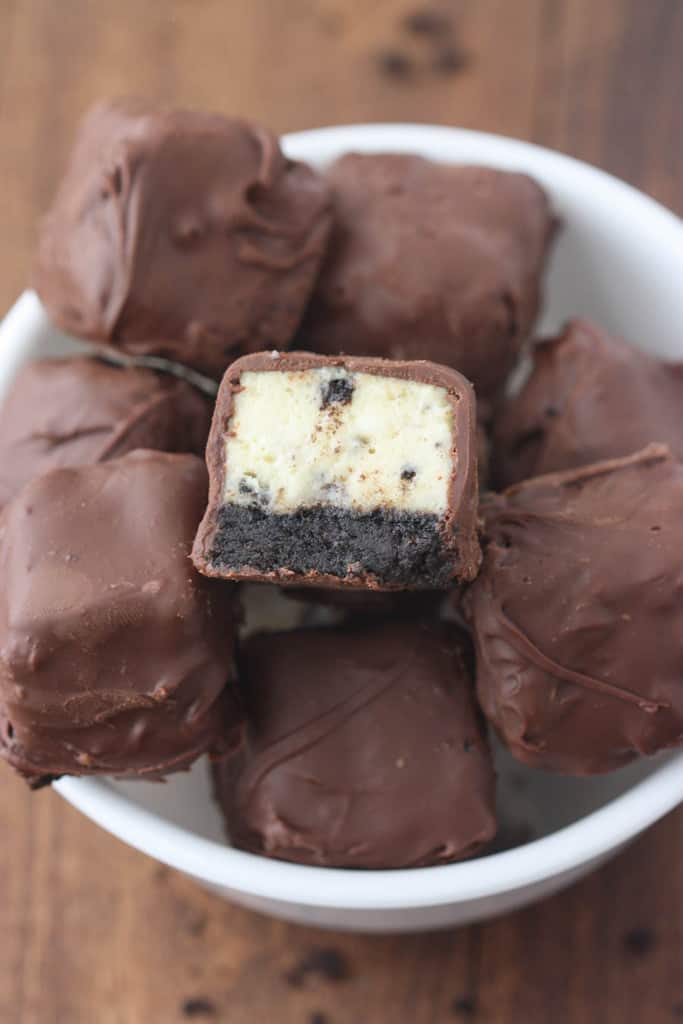 A white bowl filled with about a dozen small chocolate covered cheesecake bites with Oreo crust.
