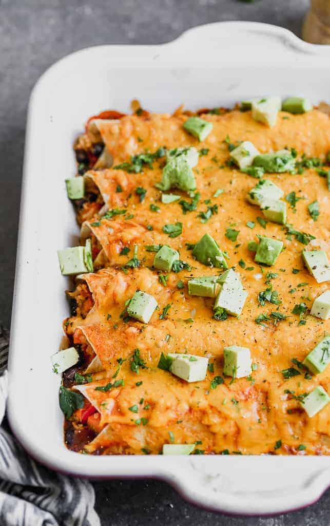 A casserole dish with vegetarian enchiladas covered in red sauce, melted cheese, and chopped avocado on top.