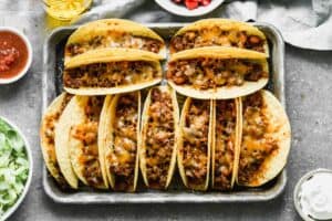 Hard shells tacos filled with a taco meat and bean mixture and topped with cheese, lined on a baking sheet and baked.