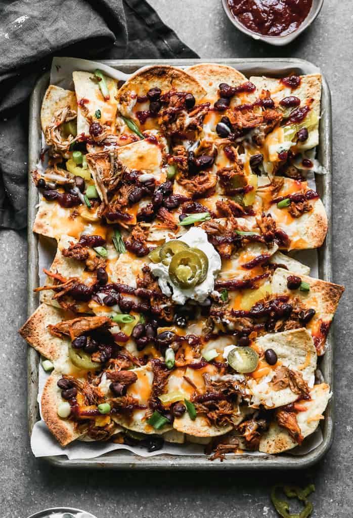 A sheet pan with pulled pork nachos on it, drizzled with BBQ sauce and scoop of sour cream in the center. 