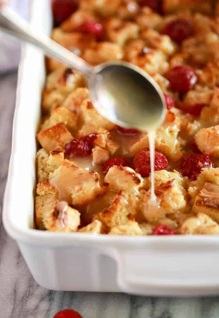 A pan of bread pudding with raspberries in it and a spoon drizzling a sauce on top.