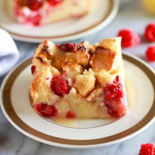 Classic Bread Pudding with raspberries in it and a vanilla cream sauce on top, served on white plate with gold trim and another serving plate in the background.