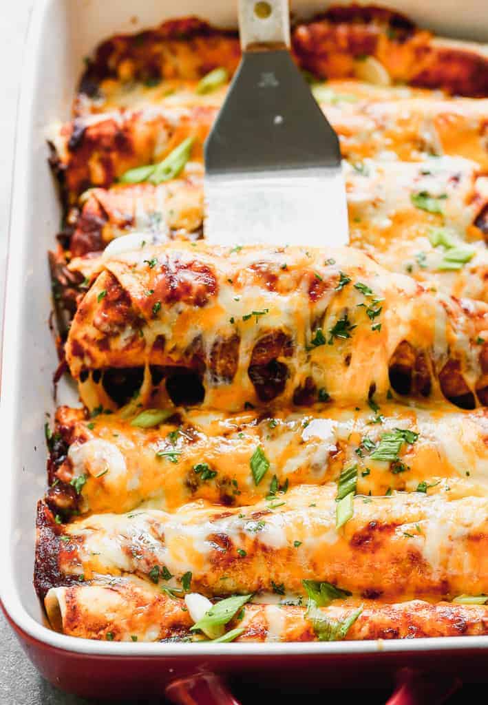 Beef enchiladas in a pan with a spatula lifting one out.