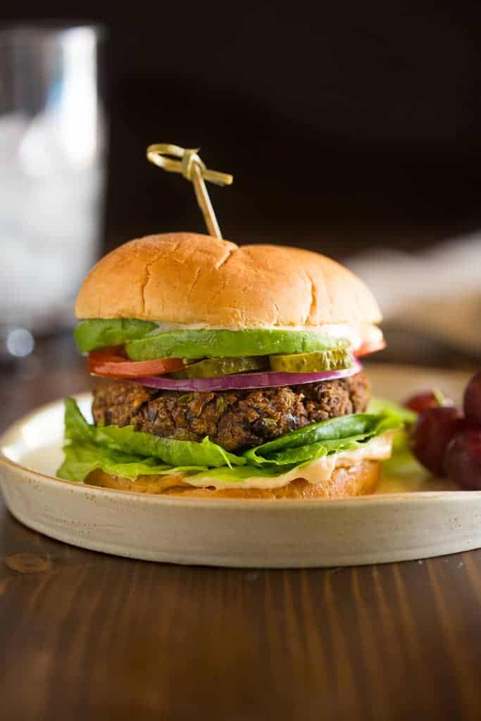 A black bean burger topped with onion, avocado, tomato pickle and lettuce with a toothpick to secure the bun and grapes served on the side.