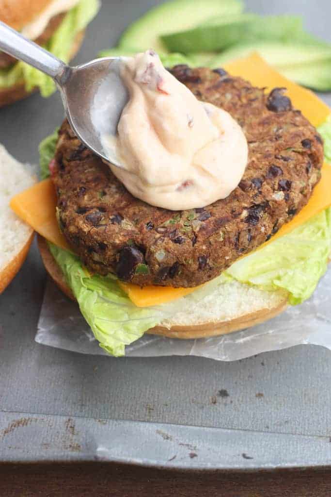 Chipotle mayo sauce being spread on top of a black bean burger patty that is sitting on cheddar cheese, lettuce, and a white bun.