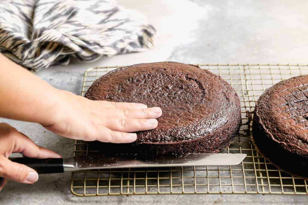 A baked round chocolate cake layer being cut in half horizontally with a serrated knife.