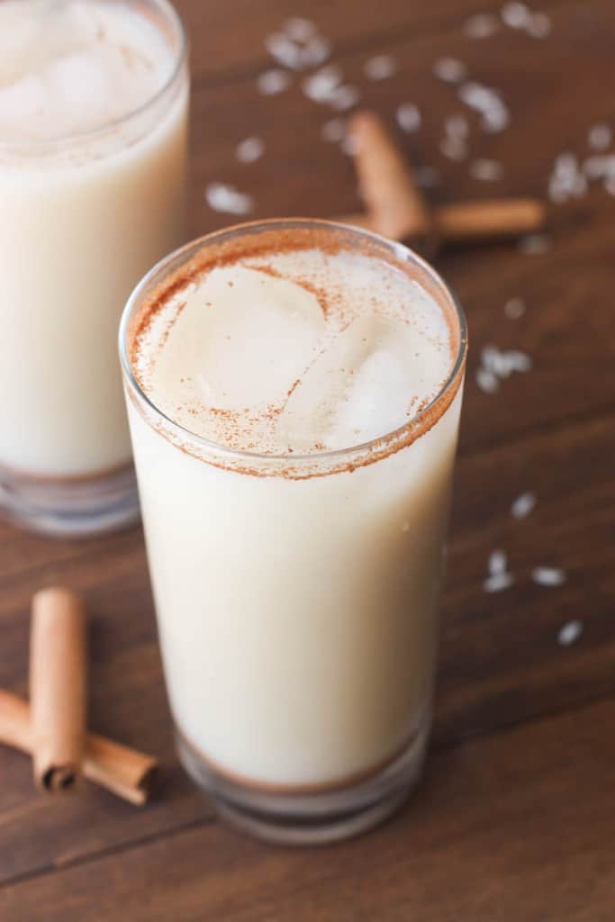 A tall, clear glass of Mexican horchata sprinkled with cinnamon and sitting on a wood table surrounded by cinnamon sticks.