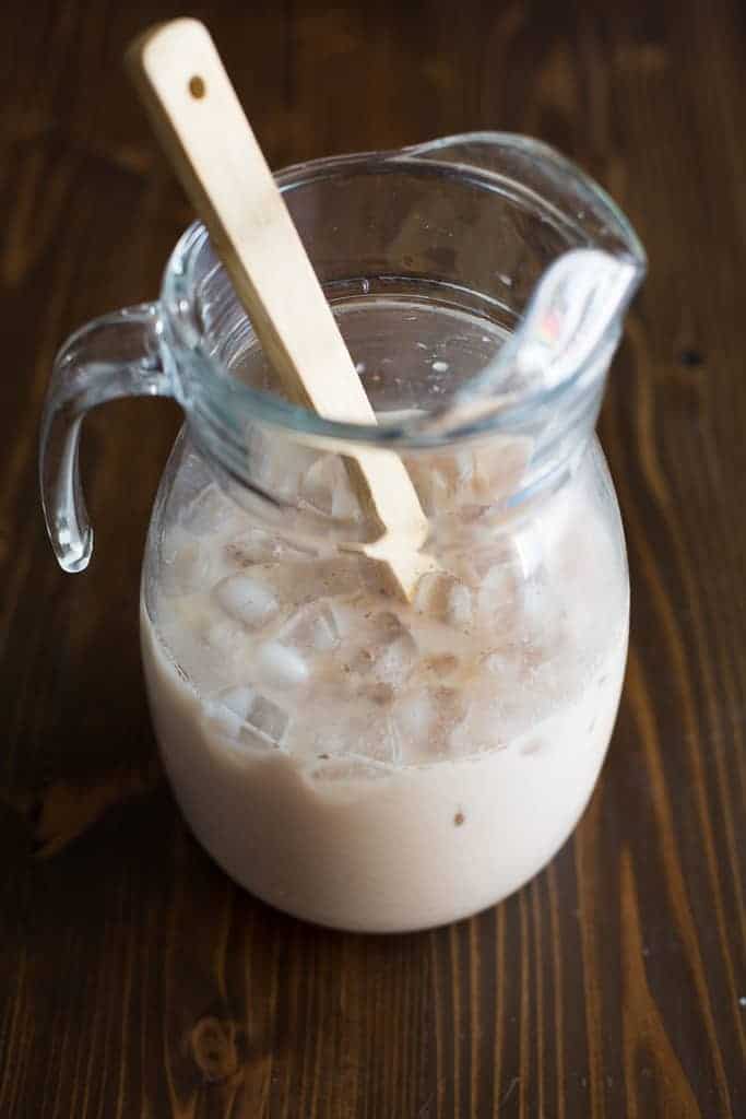 A large clear, glass pitcher with horchata, ice and a wooden spoon in it.