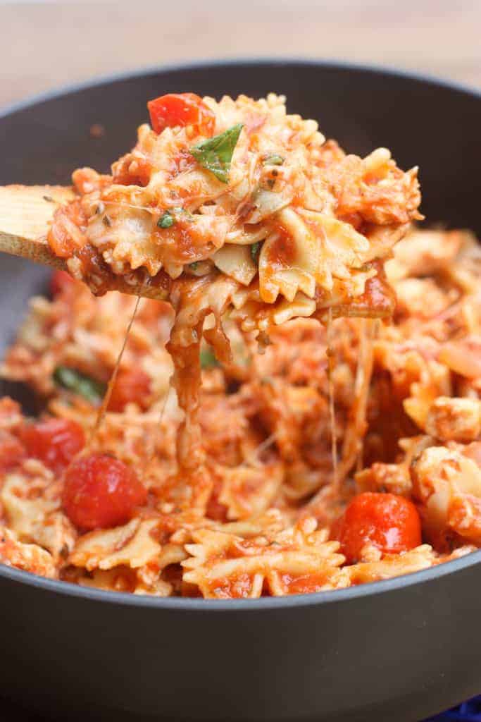 A wooden spoon scooping Chicken Mozzarella Pasta with Roasted Tomatoes out of a pan.