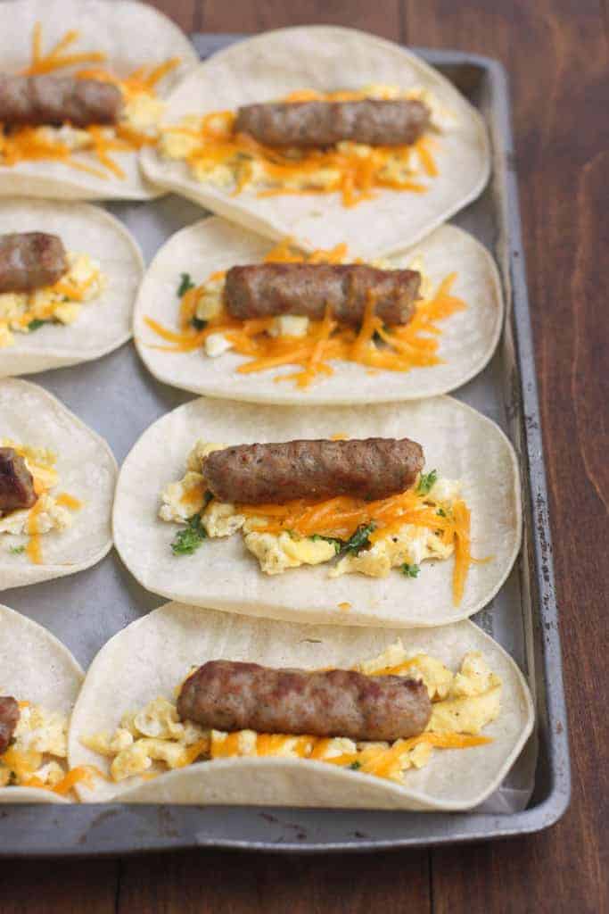 A sheet pan covered with small flour tortillas that are each topped with eggs, cheese, and a sausage link.